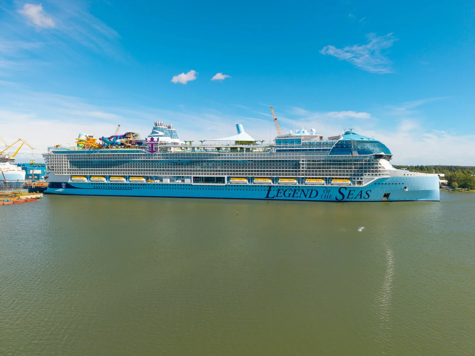 Very large new cruise ship with waterslides on top, moored at a shipyard under blue sky
