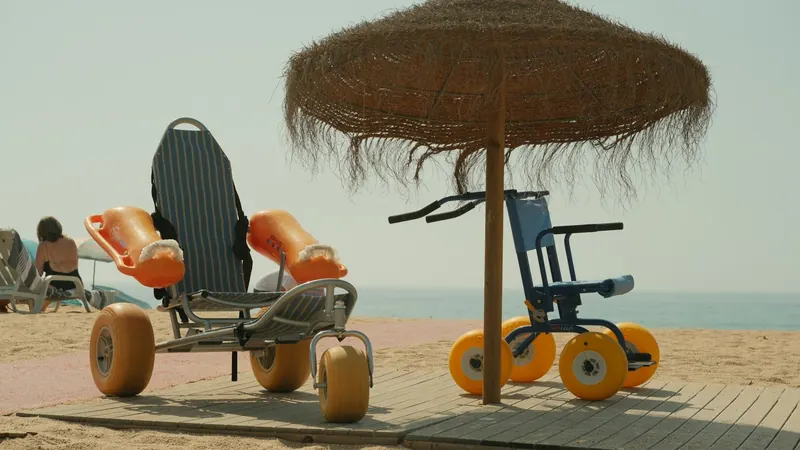 Two beach wheelchairs with wide balloon tires under a straw umbrella on the sand