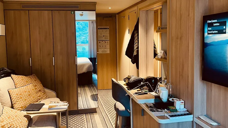 Cruise ship cabin with a sofa, desk and television, with the balcony beyond